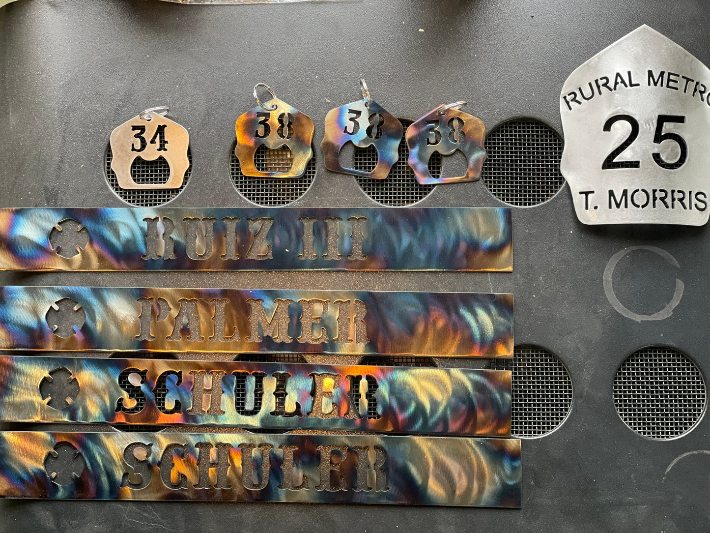 "Firefighter helmet shield keychains in various finishes, including raw steel, black powder coat, and torch patina, with personalized unit number, custom firefighter accessories for keys or gear."